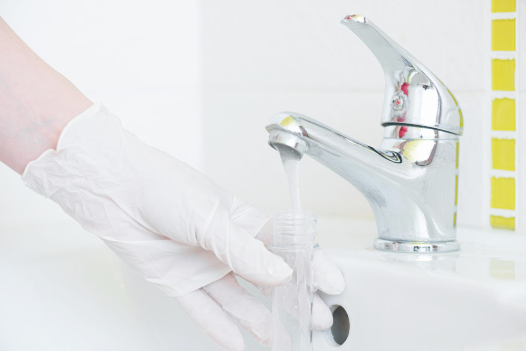 A person's hand in a white rubber glove, holding a white plastic tube under a silver bathroom faucet running water.