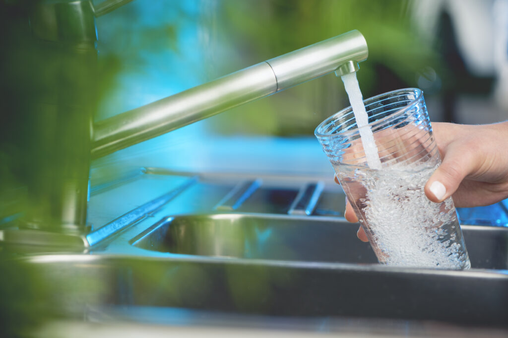 Stainless steel kitchen sink faucet flowing with clean, filtered water. Person's hand holding a drinking glass under the tap, filling it with water.